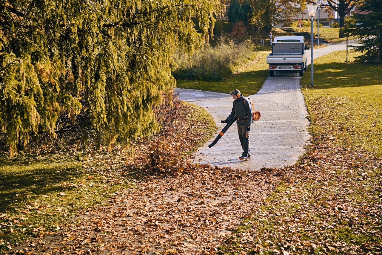 A man in work equipment using a cordless leaf blower to remove leaves from a park path